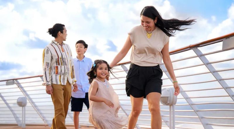 A family of four, two parents and their children, a boy and a girl, stroll along the upper deck of a cruise ship