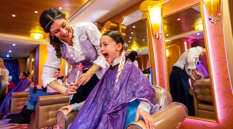 A young girl gets a royal makeover in a salon chair at the Bibbidi Bobbidi Boutique, attended by a friendly Cast Member