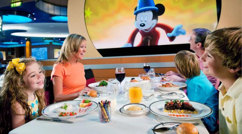 A family of five enjoys a meal together at a dining table while Mickey Mouse from 'Fantasia' plays on a nearby screen