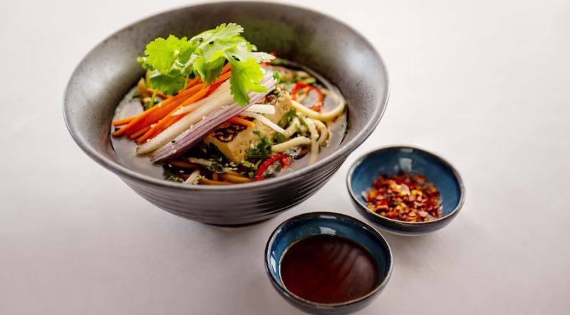 A bowl of noodle soup with tofu and fresh vegetables is served alongside two cups of sauce and a sprinkle of red chili flakes